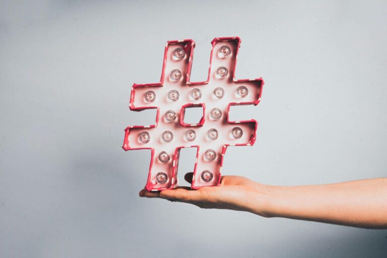 Un plan plat minimaliste avec une main tenant un grand symbole hashtag (#) lumineux et rose. Il est entouré artistiquement de fournitures de couture : bobines de fil, ciseaux de tailleur, épingles et tissu. Le texte sur l'image demande : 'HASHTAGS INSTAGRAM 2026 : FIN D'UNE ÉPOQUE OU NOUVEAU DÉPART ?'. Illustre la stratégie de référencement pour les créatrices