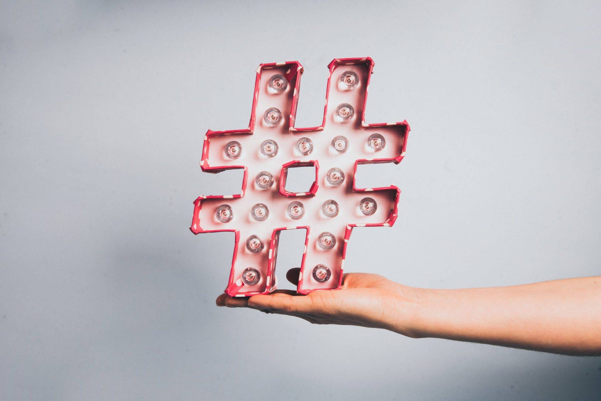 Un plan plat minimaliste avec une main tenant un grand symbole hashtag (#) lumineux et rose. Il est entouré artistiquement de fournitures de couture : bobines de fil, ciseaux de tailleur, épingles et tissu. Le texte sur l'image demande : 'HASHTAGS INSTAGRAM 2026 : FIN D'UNE ÉPOQUE OU NOUVEAU DÉPART ?'. Illustre la stratégie de référencement pour les créatrices